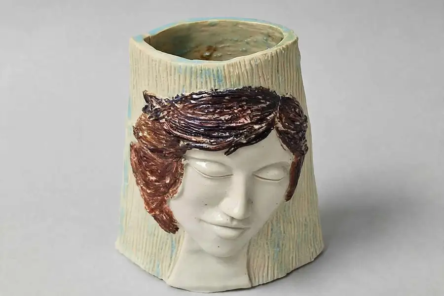 Ceramic vessel with a sculpted, closed-eyed face and cascading dark red hair